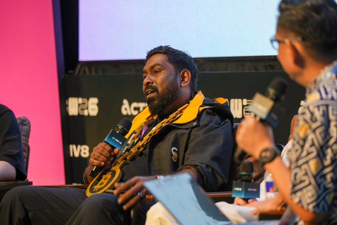 A man with a beard and a yellow scarf speaks into a microphone at a panel discussion, with branding for 'act6' and 'IVB' visible in the background.