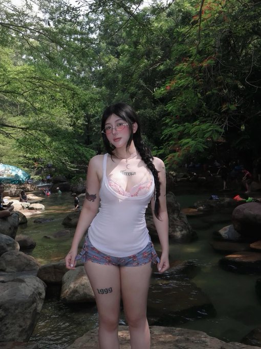 A young woman with long dark braided hair and glasses poses by a rocky stream in a lush forest, wearing a white tank top and patterned shorts, with "1999" visible on her thigh tattoo and another tattoo on her chest. The "gm" (good morning) greeting and friendly message align with the casual, nature-filled photo, inviting warm engagement from her audience.