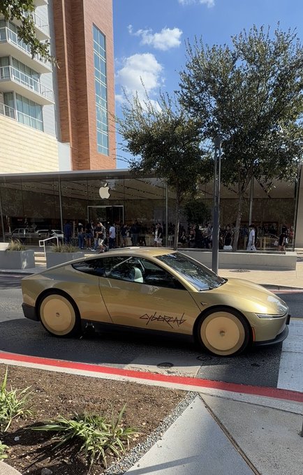 A gold/champagne colored Tesla Cybertruck parked on a street outside an Apple Store, with the word 'CYBERTRUCK' written on its side.