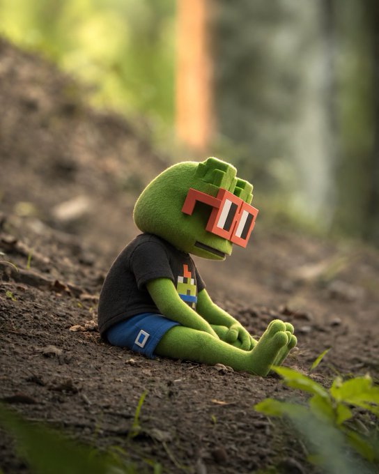 A 3D-rendered green plush toy character wearing a Minecraft-style pixelated orange glasses, black shirt, and blue shorts, sitting on dirt in a forest setting.