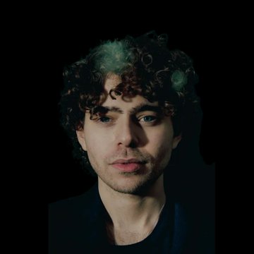 A young man with curly hair and a green-tinted highlight looks directly at the camera against a black background.

The image appears to be a portrait that does not visually connect to the accompanying post about the Polymarket phishing attack and $3.1M PUSD theft—no logos, tickers, names, or on-screen text are visible to link the photo to the news content.