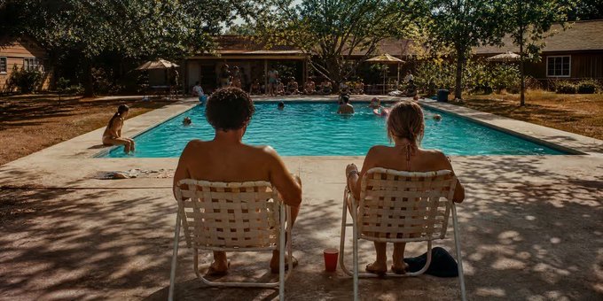 Two people sit in white lounge chairs facing a backyard swimming pool where others are swimming, with trees and a house visible in the background.
