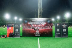 A nighttime view of a soccer field with stadium lights, featuring a banner reading 'Builders Cup' with a trophy displayed in front of it, flanked by smaller banners on either side.
