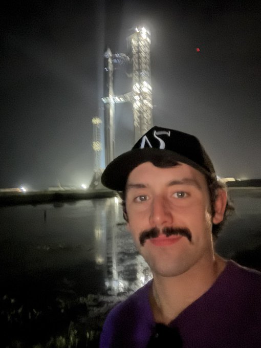 Nighttime selfie of a man wearing a black cap with mustache, taken in front of a SpaceX Starship rocket on a launch pad with bright illumination.