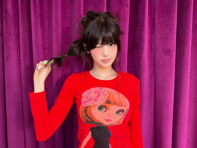 A woman with dark hair in pigtails wearing a red long-sleeved shirt with a blonde doll graphic stands against a purple velvet curtain, holding her braid.