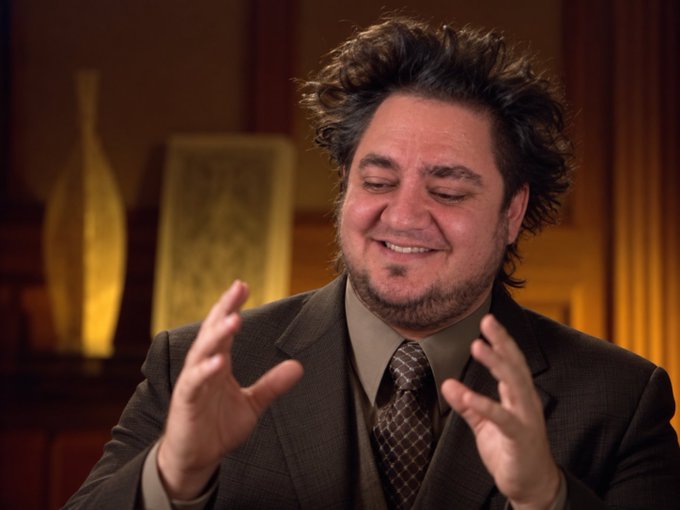 A smiling man with messy dark hair and a beard, wearing a brown suit and patterned tie, gestures with both hands raised while speaking in a warmly lit room.