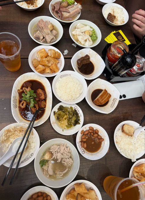 A wooden table is covered with numerous white bowls containing various Chinese dishes, including rice, soup with meat, tofu, and vegetables, along with drinks and condiment bottles.