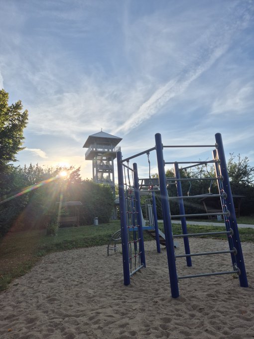 A playground with blue climbing equipment and sandy ground at sunset, with a watchtower and the sun visible in the background.