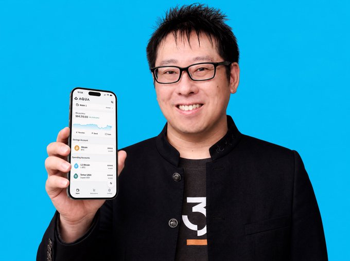 A smiling man with glasses wearing a black jacket over a t-shirt holds up a smartphone displaying the Aqua cryptocurrency app against a bright blue background.