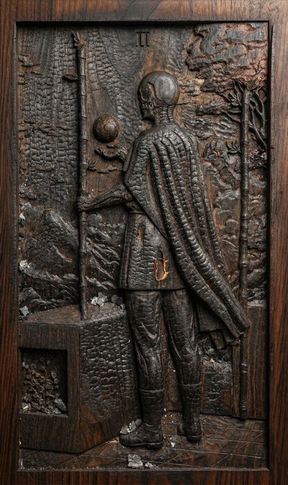 A detailed wood-carved relief sculpture depicting a hooded figure holding a staff and orb, facing a stone wall, with Roman numeral 'II' visible at the top and intricate textures throughout the dark wooden panel.
