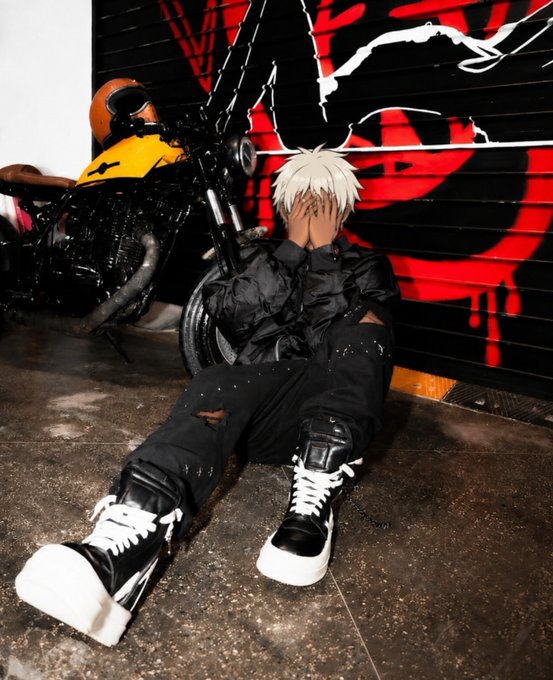 A cosplayer with white hair sits on the floor covering their face with their hands, wearing a black outfit and high-top sneakers, posed next to a yellow and black motorcycle against a graffiti wall.