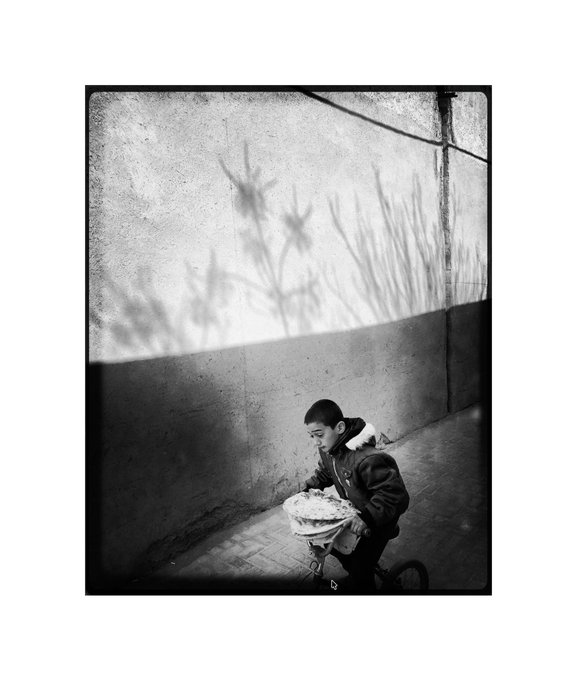 A black-and-white photograph shows a young boy in a hooded jacket riding a bicycle while carrying flatbreads, with the shadow of flowering plants cast on the wall behind him.