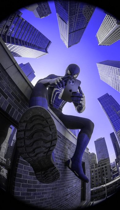 A cosplayer dressed as Spider-Man sits perched on a brick ledge holding a tablet, captured with a dramatic fisheye lens that makes surrounding skyscrapers tower above against a vivid purple-blue sky. The tech-savvy pose with the device playfully ties to the post's promotional hashtags #InterLink, #ITLG, and #ITL, suggesting the hero is geared up for a digital milestone or announcement.