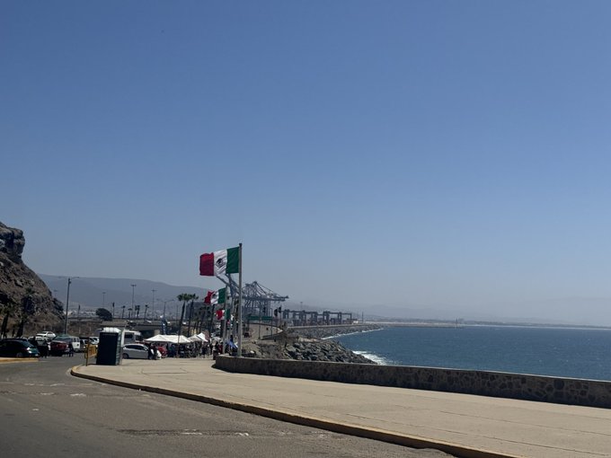 A scenic coastal view showing a walkway with Mexican flags, a port with shipping cranes in the distance, and the ocean under a clear blue sky.