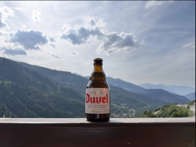 A bottle of Duvel Belgian beer sitting on a railing with a scenic mountain landscape and cloudy sky in the background.