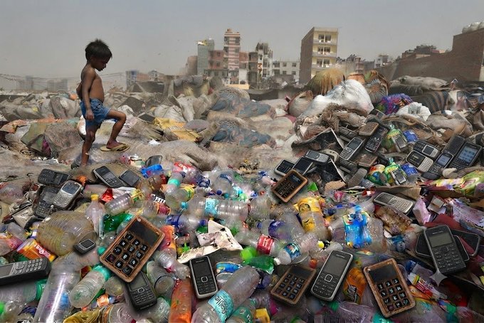 A child walking on a large pile of e-waste including discarded mobile phones, plastic bottles, and electronic components in what appears to be a developing area.