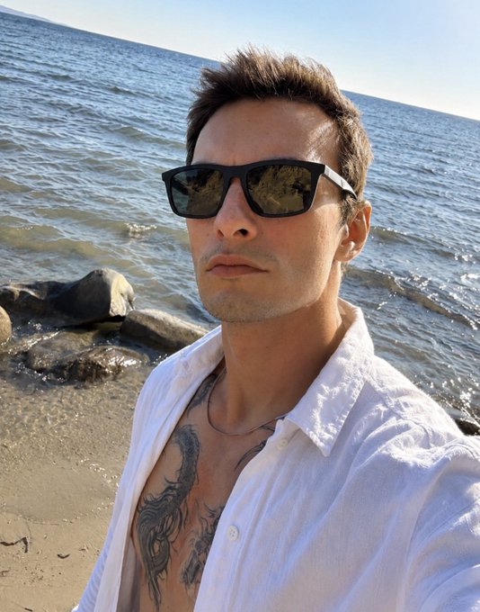 A man takes a selfie on a beach, wearing black sunglasses and an unbuttoned white linen shirt that reveals a chest tattoo, with the ocean and rocks visible behind him.
