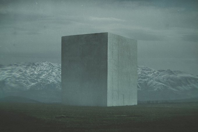 A large monolithic concrete cube sits isolated in an empty grassy field, with snow-capped mountains and a cloudy, moonlit sky in the background.