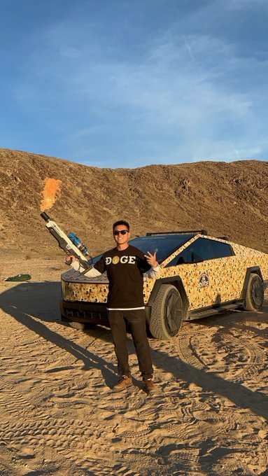 A man wearing sunglasses and a black 'DOGE' shirt stands in front of a custom desert-camo-wrapped Tesla Cybertruck in a desert landscape, holding what appears to be a flamethrower.