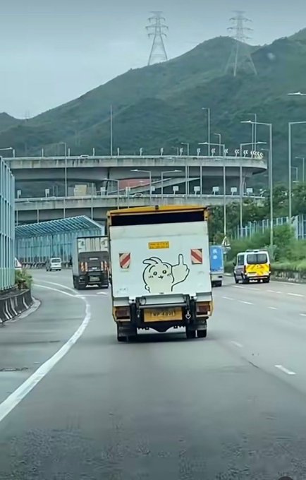 A box truck with a cartoon dog logo on its rear doors drives down a highway, passing under an overpass with power line towers visible against the mountainous backdrop.