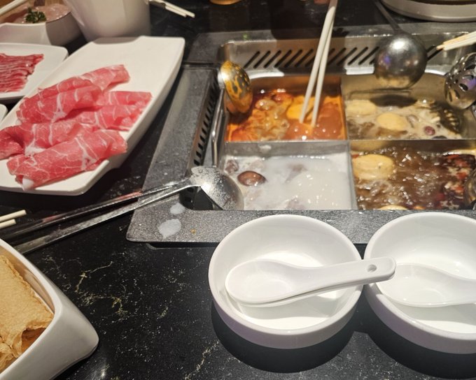 A four-section hot pot meal with various broths, raw sliced meat on a plate, and empty serving bowls ready for diners.