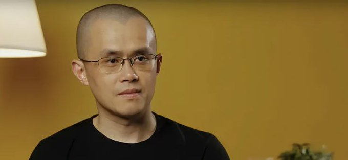 A bald man wearing glasses and a black t-shirt sits in a softly lit room with a lamp on one side and a plant in the background.