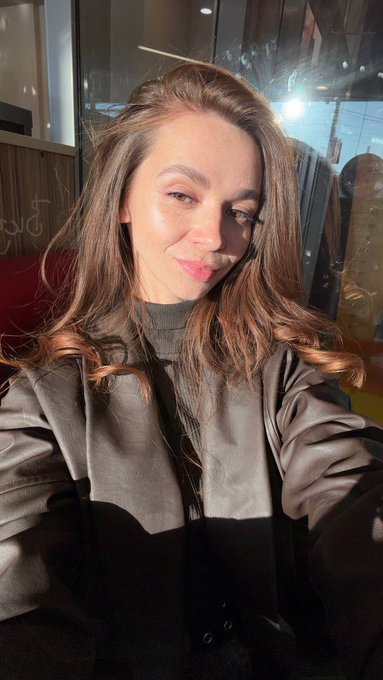 A selfie of a smiling woman with long brown hair wearing a dark coat, illuminated by sunlight.