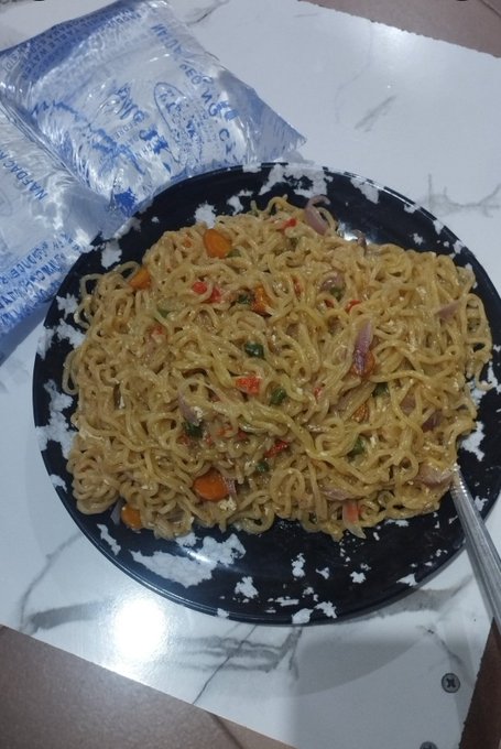 A plate of stir-fried noodles with vegetables and meat, served on a black plate on a white marble surface with a plastic water bottle beside it.