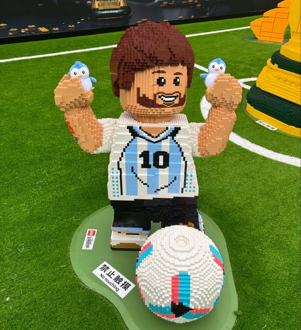 The image features a Lego man figure holding two penguins, with a soccer ball placed between them. The surrounding area includes a green field and other Lego figures scattered around. The main focus of the image is the Lego man figure holding the soccer ball in his hands.