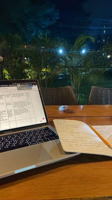 An open MacBook Air displaying a spreadsheet and a handwritten notebook with a pencil sit on a wooden table outdoors at night, with palm plants and lights visible in the background.