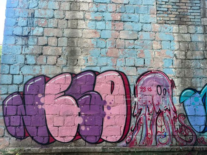 A weathered brick wall features colorful graffiti, including pink and purple bubble letters and a pink jellyfish-like figure with "XXX 00" text, serving as a vibrant street art backdrop for a crypto whitelist announcement tagging several usernames including @_alphavault.