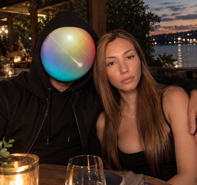 A woman with long brown hair in a black top sitting at an outdoor restaurant table at dusk with a waterfront view, next to a person in a black hoodie whose face has been replaced with a colorful iridescent sphere.