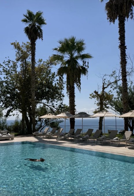 A person swims in a resort pool surrounded by palm trees, white lounge chairs, and umbrellas with the ocean visible in the background.