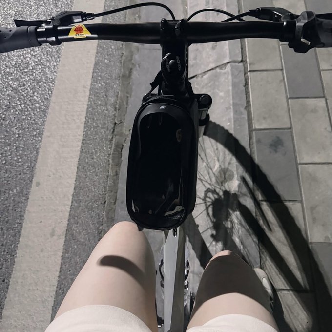 A first-person view from a bicycle, showing the handlebars, a black bag attached to the front, and the rider's legs over a paved path.