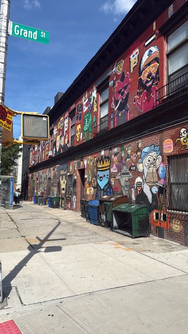 A street scene at 'Grand St' showing a building facade covered in colorful murals and cartoon character artwork, with green dumpsters lined up on the sidewalk.