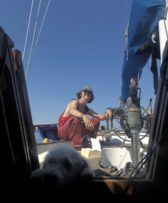 A woman wearing a red outfit and a grey cap smiles while crouching on the deck of a sailboat under a clear blue sky, with a dog visible in the foreground.