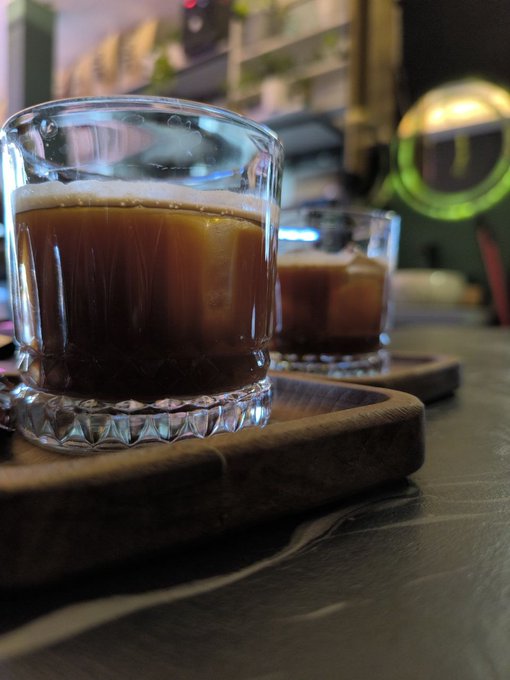 Two glasses of iced coffee sit on a wooden serving board at a cafe, with a blurred espresso machine glowing in the background. The cozy setting complements the post's weekend vibes, capturing a relaxing moment spent at a cafe with university friends after a day of hiking.