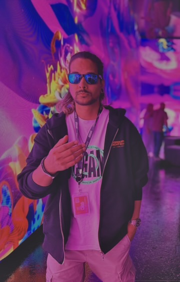 A young man wearing sunglasses, a hoodie, and a "GN" lanyard poses confidently in a vibrant, neon-lit immersive art space, with the "GN" branding tying directly to the "GN Apes" post theme. The colorful psychedelic backdrop and his lanyard reinforce the connection to the GN Apes community.