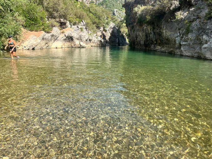 A woman wades into a crystal-clear turquoise pool surrounded by rocky cliffs and green vegetation at a secluded natural swimming hole.