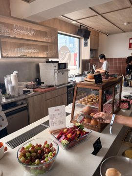 A photo of a bakery or cafe counter with a bowl of grapes and plums in the foreground, pastries in a display case, and staff working in the background.