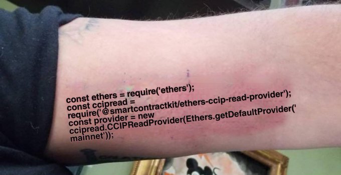 A tattoo of JavaScript code on someone's forearm, importing the `ethers` library and `@smartcontractkit/ethers-ccip-read-provider` to instantiate a `CCIPReadProvider`, humorously representing the user's dedication to Chainlink's CCIP-Read technology as they muse over a hypothetical merger between Chainlink and ENS to complete the Chainlink Stack's name services.