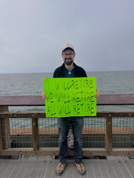 A man standing on a pier by the ocean holds a bright green sign with handwritten text reading 'WE WILL ARE TIRE / WE WILL YET TIRE / ALL WILL GET TIRE'.