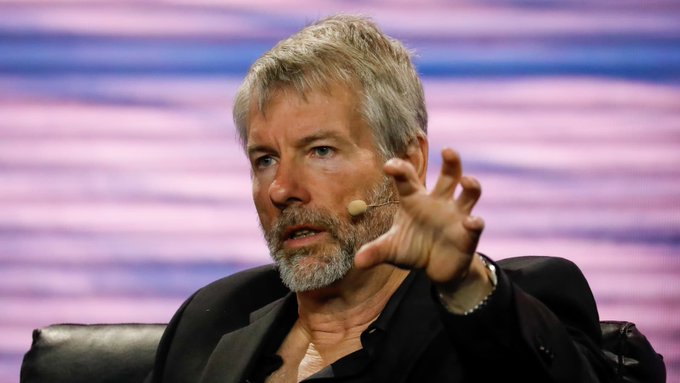 A man with gray hair and beard, wearing a black shirt, speaks while gesturing with his hand at what appears to be a conference or panel discussion.