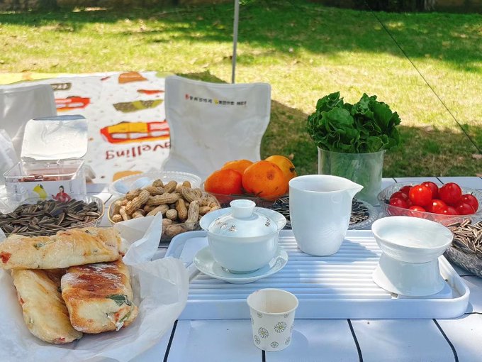 An outdoor picnic table set with various foods including pastries, peanuts, sunflower seeds, persimmons, cherry tomatoes, sunflower greens, and a traditional tea set with a white gaiwan and pitcher.