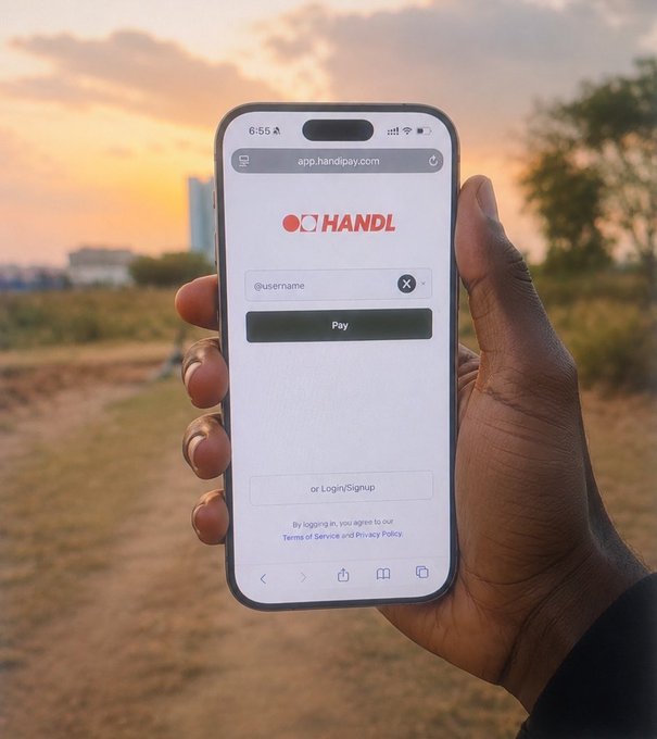 A hand holding a smartphone outdoors at sunset, displaying the app.handipay.com login screen for 'HANDL' with a prompt to enter a username to pay.