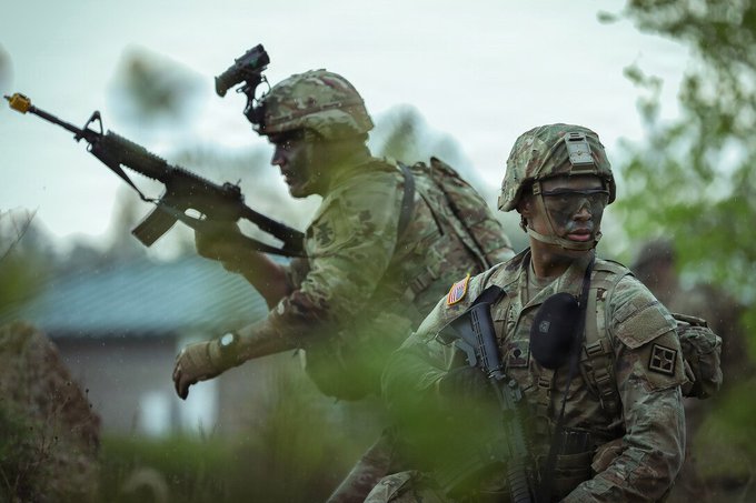 Two soldiers in camouflage uniforms and helmets with American flag patches are seen in a field, one aiming an M4 rifle while the other holds a radio, suggesting a combat or training scenario.