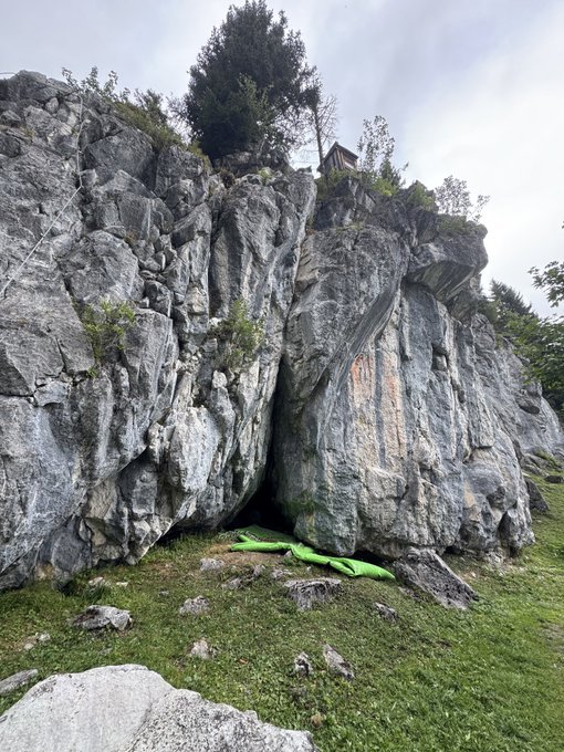 A rocky cliff face with green climbing crash pads laid out at the base on the grass, indicating a bouldering site.