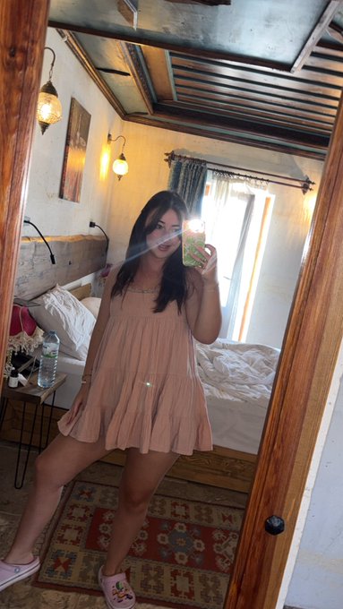 Mirror selfie of a woman in a pink dress standing in a rustic room with wooden beams and a bed.