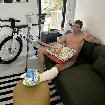 A shirtless man with a bandaged cast on his leg gives a thumbs up while holding a remote and a pizza box, with the partial text "ANVO" visible in the background. The image humorously illustrates the post's claim about Joe Nakamoto's high ankle sprain, suggesting he's recovering comfortably at home after his hiking mishap in designer boots.