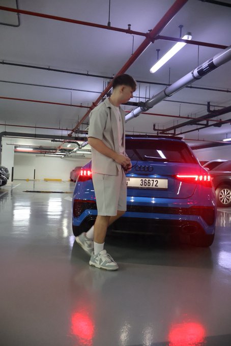 A young man in a casual beige outfit poses confidently next to a sleek blue Audi SUV with license plate "36672" in a parking garage, embodying the "get money" lifestyle through his trendy attire and luxury vehicle.